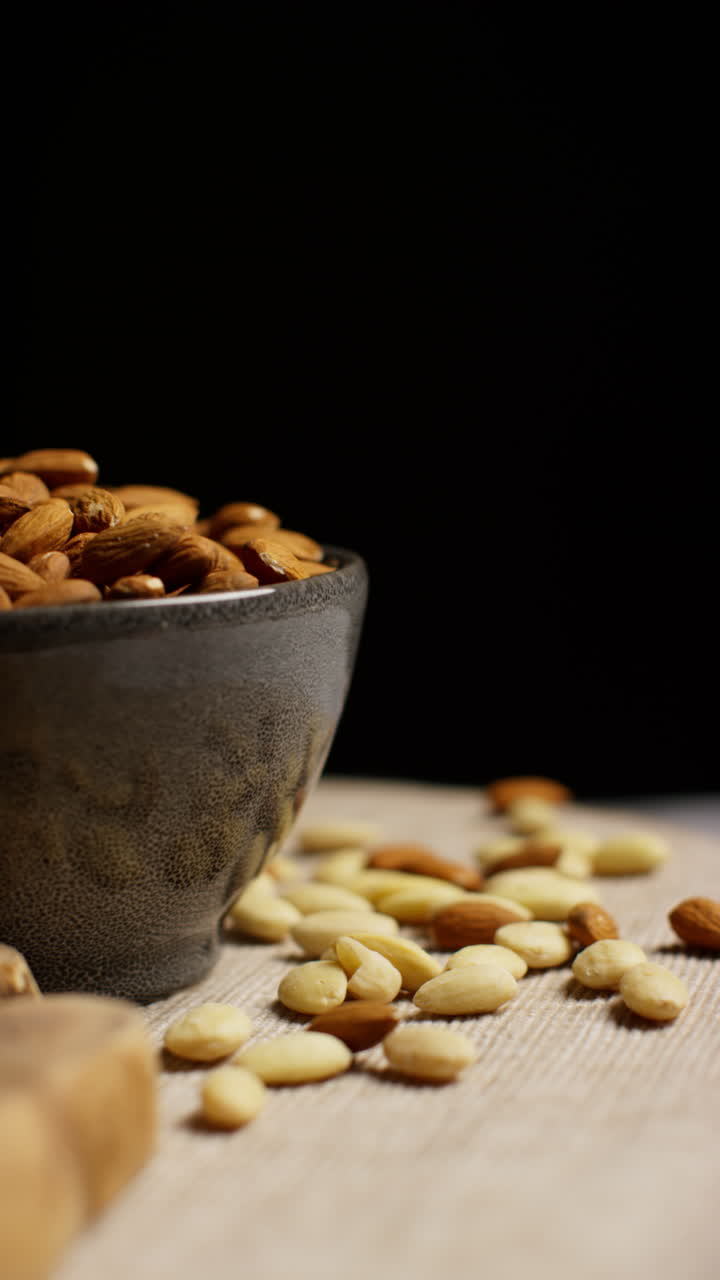 Vertical Video Close Up Studio Shot Of Bowl Of Raw And Blanched Almonds Revolving Against Black Studio Background