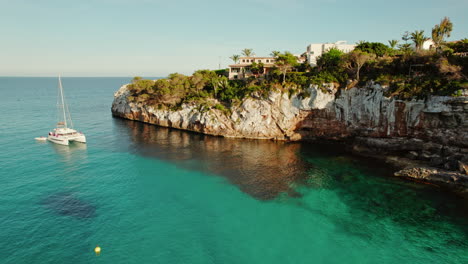 Catamaran-Boat-Floating-In-Crystal-Clear-Water-Next-To-Houses-On-Cliff-In-Cala-Llombards,-Mallorca,-Spain