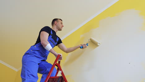 Professional craftsman painting walls indoors. Young male worker in blue overalls standing on a building ladder and use paint roller. View from below.