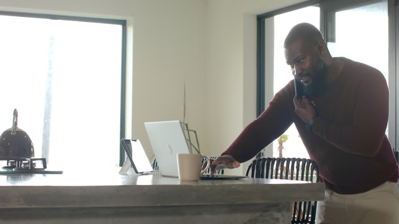 African-American man in kitchen taking smartphone call using laptop and mug, copy space