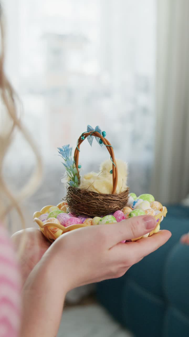 Child sits on a blue sofa, holding a miniature Easter basket. She wears pink pants and a yellow shirt. Her pigtails bounce as she looks curiously at the camera. The basket contains colorful eggs