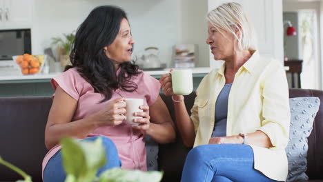 happy diverse senior women with coffee, discussing on sofa in sunny living room, slow motion