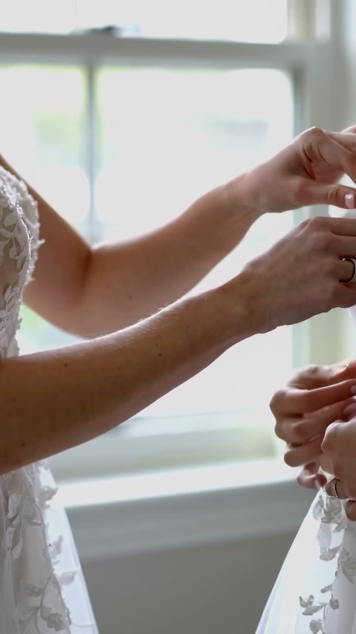 Vertical video: Backlit by window helper fastening bride gown, securing hooks and lace before vows