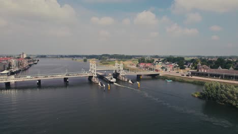 Elevated-bridge-over-a-calm-river-with-moving-boats,-houses-in-the-background,-sunny-day,-aerial-view