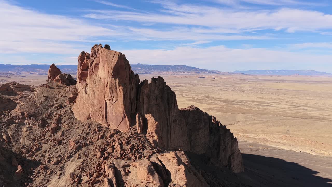 Il drone si alza per rivelare le ripide cime rocciose frastagliate e le guglie vulcaniche di un monolite montuoso all'interno di un vasto paesaggio desertico arido sotto un cielo blu con nuvole filiformi. Perfetto per ambientazioni fantasy.