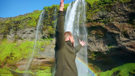 hombre posando alegremente para la cámara con un arco iris en la cascada seljalandsfoss en la región sur de islandia