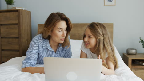 close-up view of happy young woman lying with her lovely daughter watching something on laptop lying on the bed in the morning