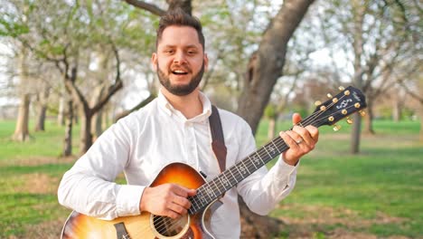 Adult-man-and-musician,-singer,-guitarist,-and-artist-is-walking-through-a-park-singing-a-song-and-strumming-his-guitar