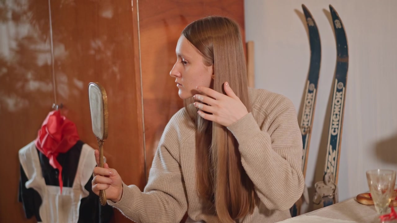 Donna che acconcia i capelli lunghi tenendo uno specchio, sistemando le ciocche mentre guarda un manichino vintage e sci sullo sfondo, interno caldo, atmosfera da camerino di moda, movimento di capelli lisci e lucidi, riflettente.