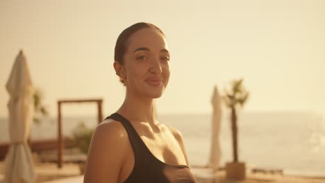 Portrait-of-a-brunette-girl-in-a-black-top-who-folds-her-arms-on-her-chest,-looks-at-the-camera-and-smiles-against-the-backdrop-of-a-sunny-beach-with-palm-trees