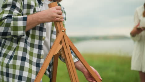 a man in a plaid shirt is setting up a wooden tripod in a grassy field. the close-up shot focuses on the man's hands and the tripod, while in the blurred background, a woman in a white dress stands