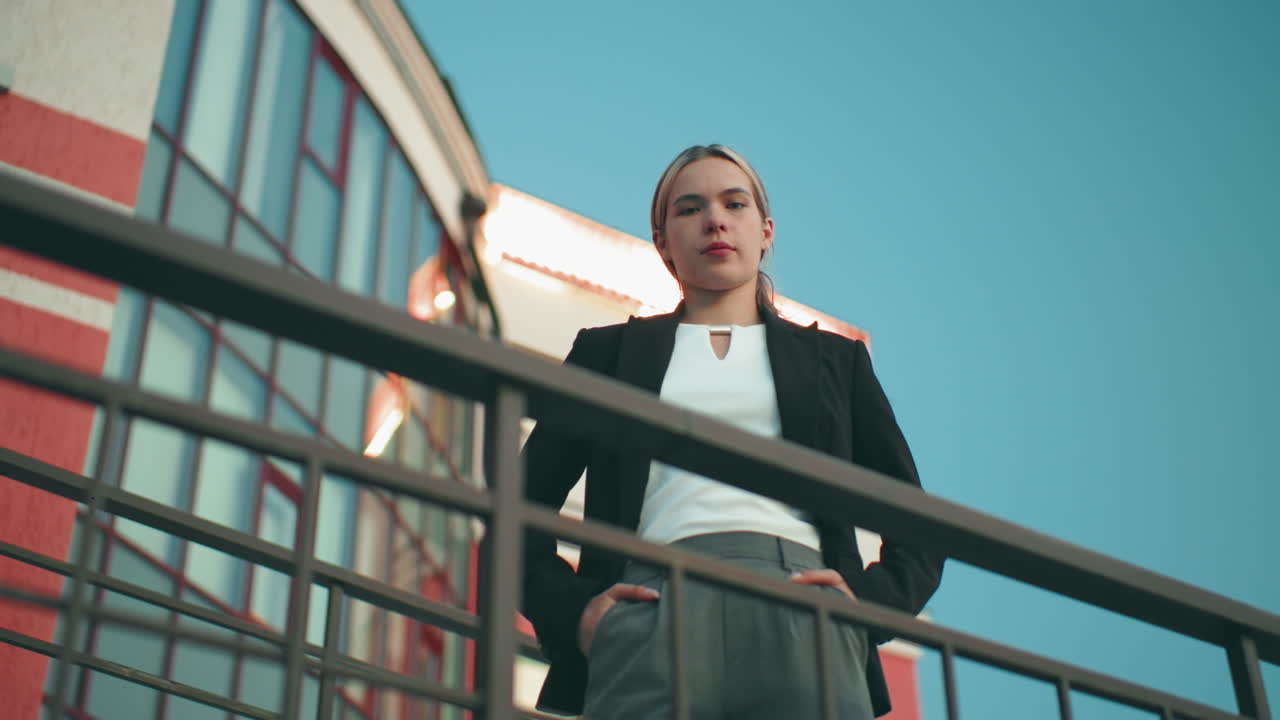 Down top view of woman confidently posing in front of camera with one hand in pocket outside modern building with reflective glass and display light blinking on roof top under clear blue sky