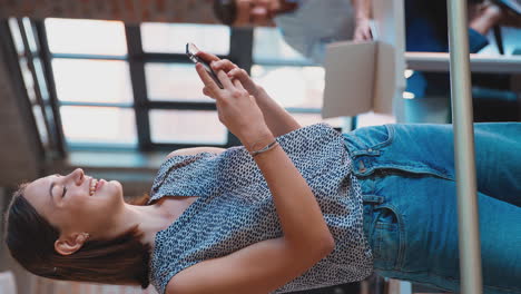 video vertical de una mujer de negocios sonriente de pie en una oficina ocupada enviando mensajes en el teléfono móvil
