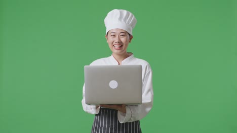 mujer chef asiática usando una computadora portátil y apuntando a un lado mientras está de pie en el estudio de fondo de la pantalla verde
