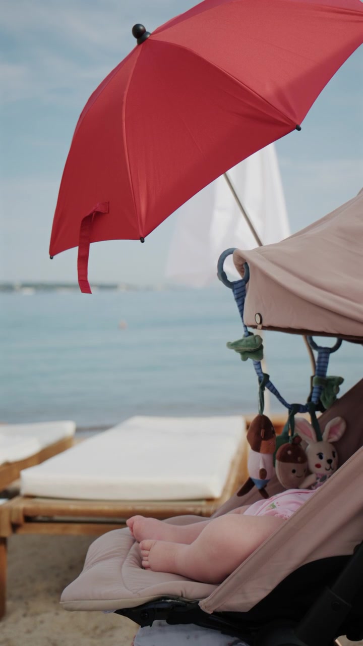 Bambino seduto nel passeggino sotto un ombrellone da spiaggia rosso brillante in riva al mare.