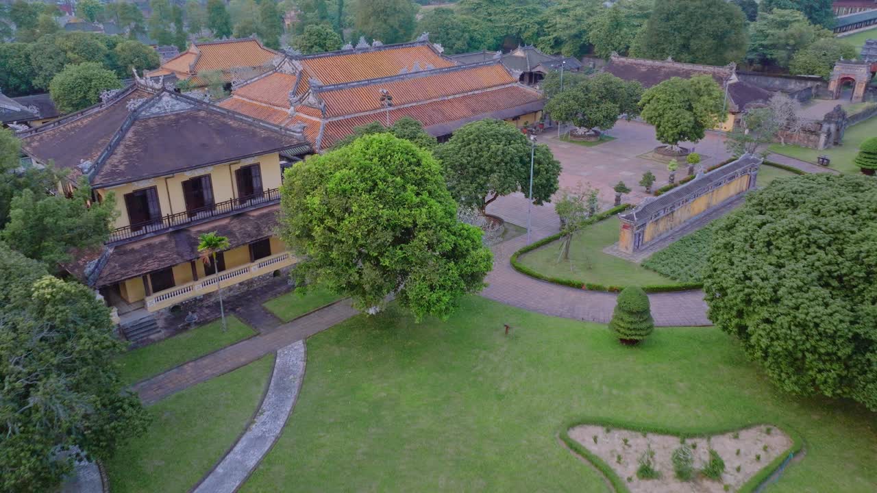 This footage captures an aerial view of the Hue Imperial City in Vietnam, showcasing its impressive traditional architecture with yellow walls and red tiled roofs, surrounded by lush green trees.