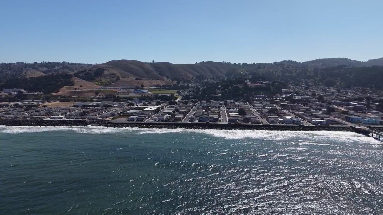 Video de dron deslizándose sobre la costa de Pacifica con la luz del sol matutina iluminando las olas del océano, el icónico muelle, el barrio costero y las ondulantes colinas de California con gran detalle. Toma con efecto dolly zoom.