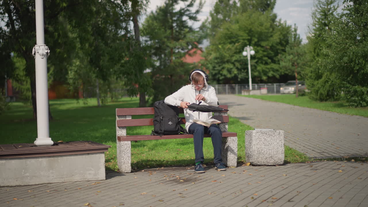 공원 벤치에 앉아 우쿨렐레를 조율하며 흐린 하늘 아래 우산을 펼치고 있는 백인 남성, 옆에는 가방과 돌 블록이 있고, 배경에는 가로등과 나무가 늘어선 통로가 보이는 생각에 잠긴 거리의 악사
