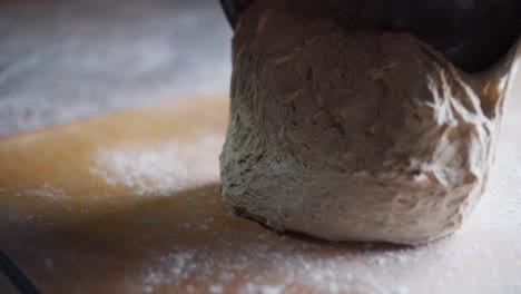 un lote suave y esponjoso de masa recién levantada siendo raspado de un cuenco de metal en una mesa de madera de la cocina, filmado como una toma en cámara lenta de primer plano