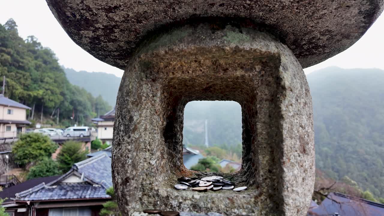 Japanese coins offered as donations rest on a stone lantern, with a waterfall and traditional houses in the background