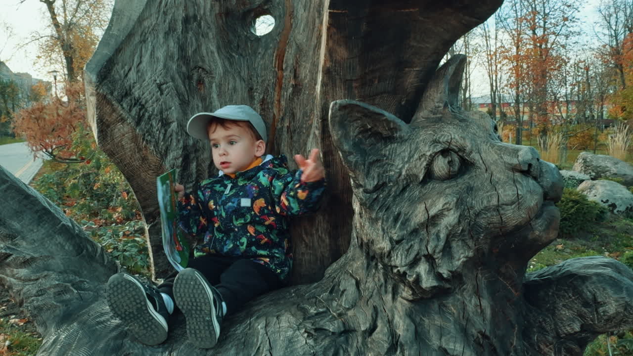 Lovely little kid sitting on the wooden cat sculpture and touching its ear. Baby boy with book outdoors.