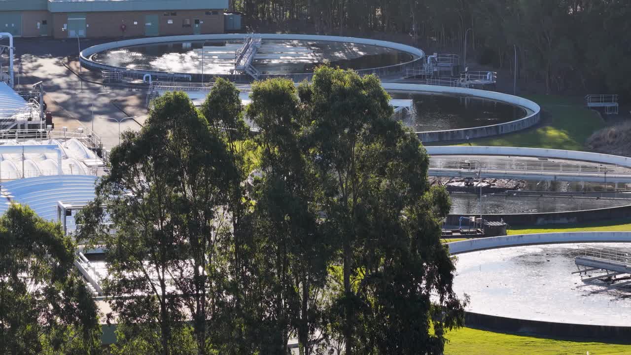Drone ascends over a modern wastewater treatment facility at sunset, revealing circular settling tanks, green trees, and industrial buildings in soft natural light