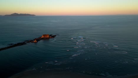 Sobrevuelo-Aéreo-Pequeñas-Olas-Del-Océano-Pacífico-Con-Rocas-Sobresalientes-Y-Hermosa-Puesta-De-Sol-Colorida-En-El-Horizonte-En-Nueva-Zelanda