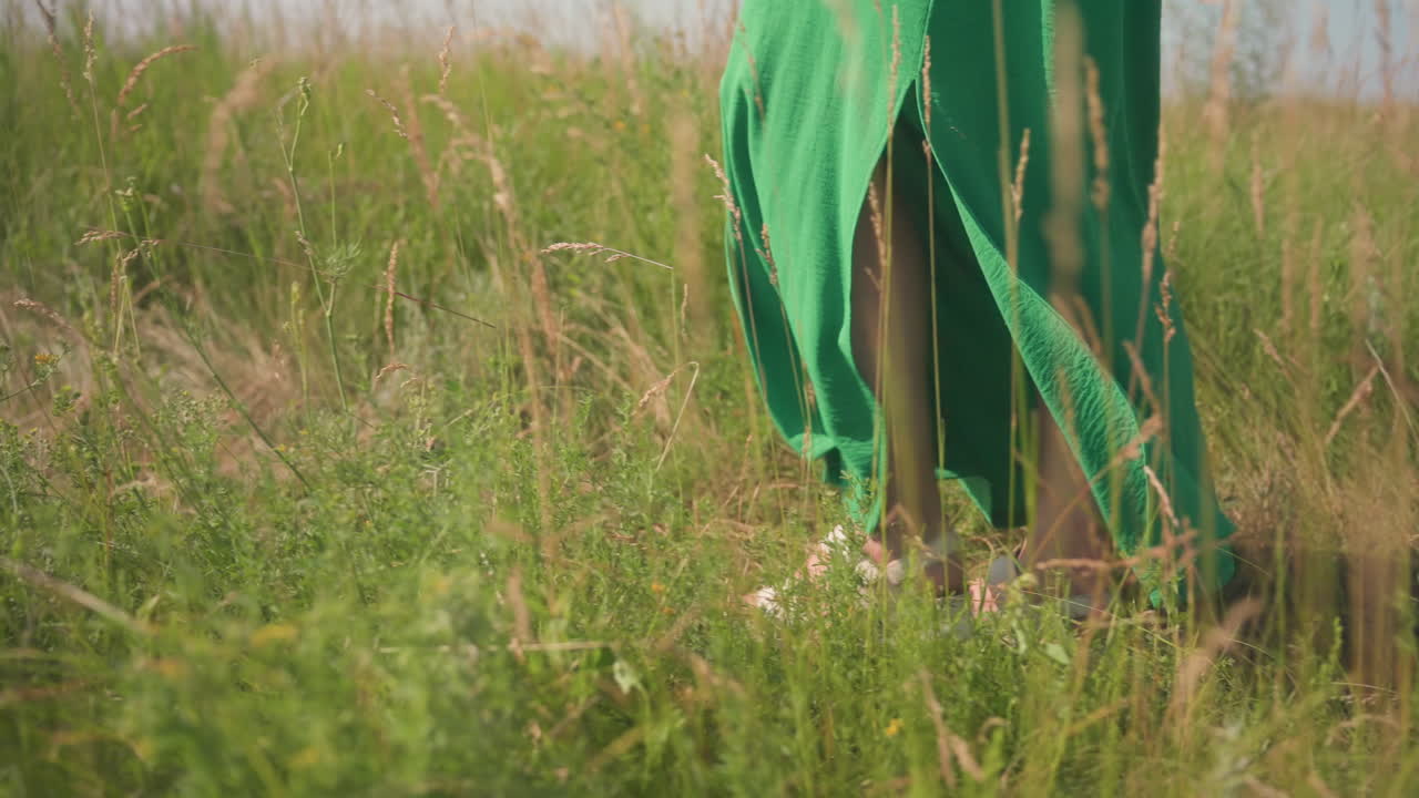 Closeup side view of girl leg in flowing green dress stepping through tall grassy field, capturing graceful movement and gentle breeze under bright summer sun