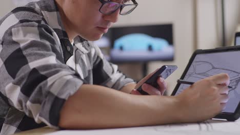 close up of asian male looking at a smartphone and drawing on a tablet while working on a car design sketch on table in the studio