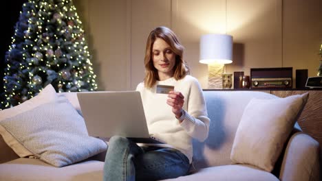 cheerful smiling beautiful woman buying on internet xmas presents paying with credit card using laptop at cozy decorated home