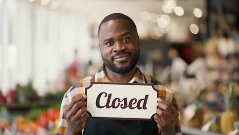 portrait of a black-skinned man in a checkered shirt and black apron holding a sign with the close sign in a supermarket. video filmed in high quality