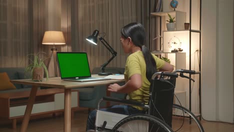 asian kid girl sitting in a wheelchair using laptop computer with mock up green screen and pain her back at home