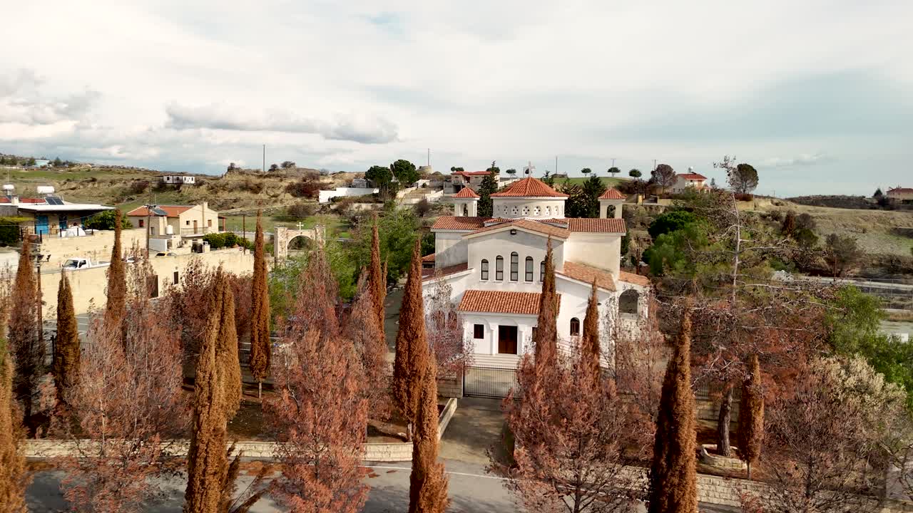 Una chiesa si erge in una zona rurale di Cipro con un tetto rosso. Il paesaggio include colline e alberi. Il cielo è nuvoloso con campi aperti sullo sfondo.