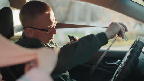 a man wearing a green jacket is seated in a car, captured in the moment of buckling his seat belt while holding a phone in his hand