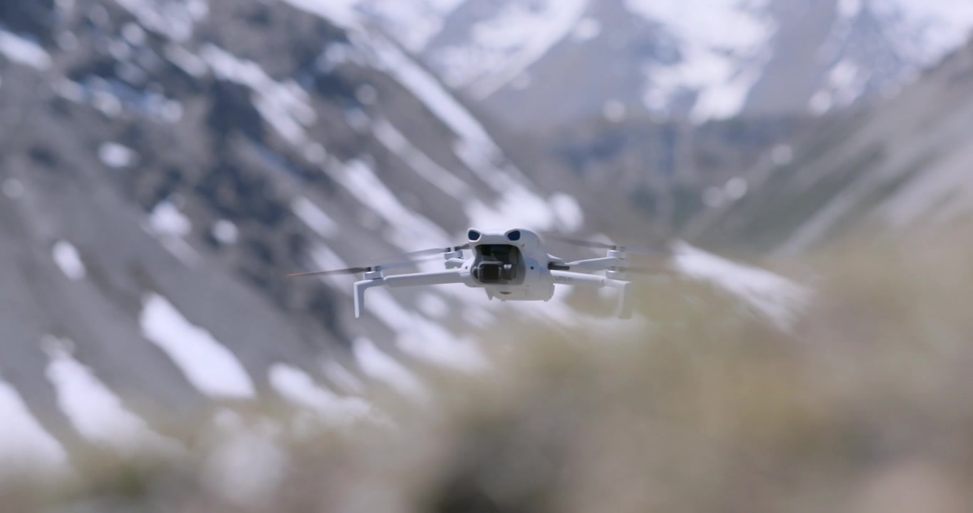 Close up of a small white drone flying low over dry grass with a snow covered mountain in the blurred background