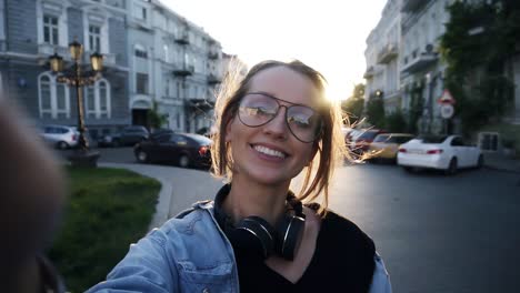 A-young-girl-in-glasses-poses-with-a-camera-on-her-outstretched-hand.-Smiling,-happy,-shows-a-hand-gesture-"Cool".-Evening-dusk