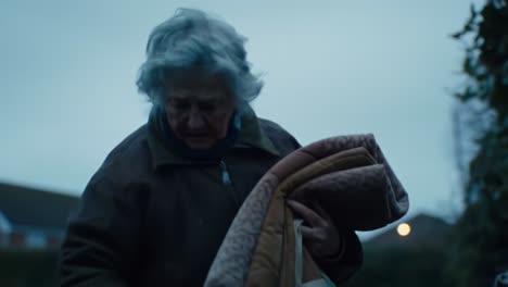 an elderly woman organizes blankets into her bag as dusk settles over a quiet rural area. she moves with care, showing tenderness in her task amidst the fading light of the day.