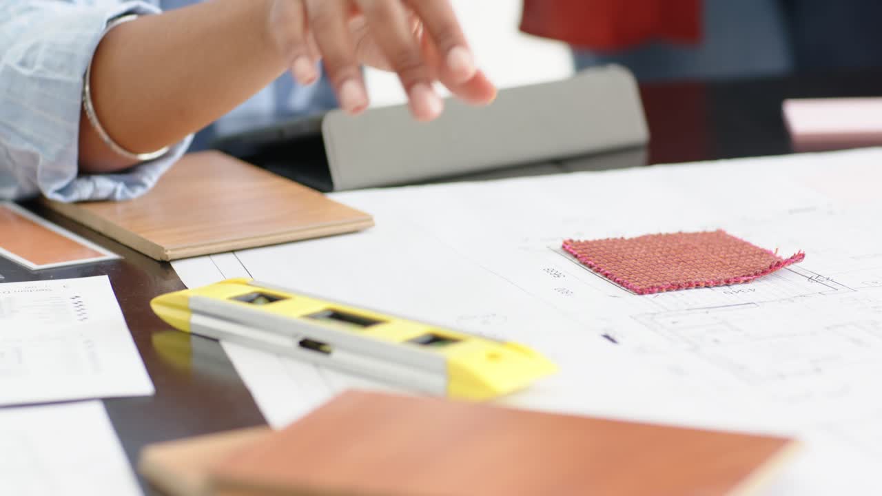 Diverse female coworkers reviewing in studio, one woman sliding level across plans near red swatch