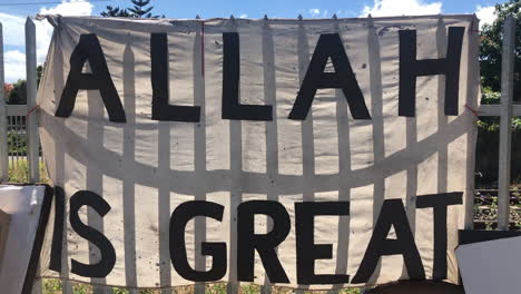 hand held shot of ragged white banner with black coloured religious text allah is great tied on a steel fence in poor urban area on a sunny day