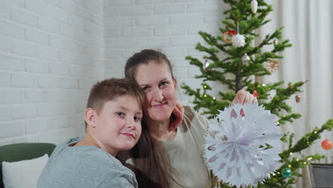 boy holding mother closely while smiling as she lifts intricate paper craft decoration near christmas tree decorated with lights indoors, joyful bonding moment captured with cozy home background