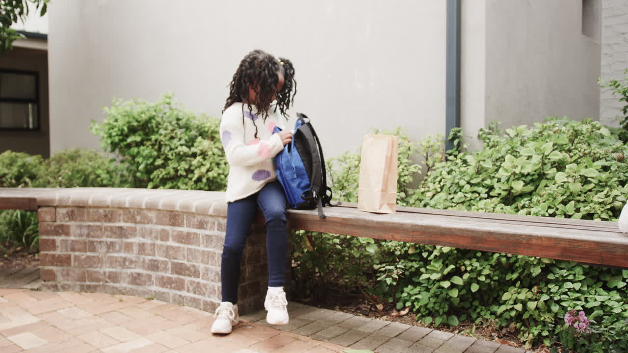 Young girl sitting on bench, opening backpack near school, enjoying break, copy space