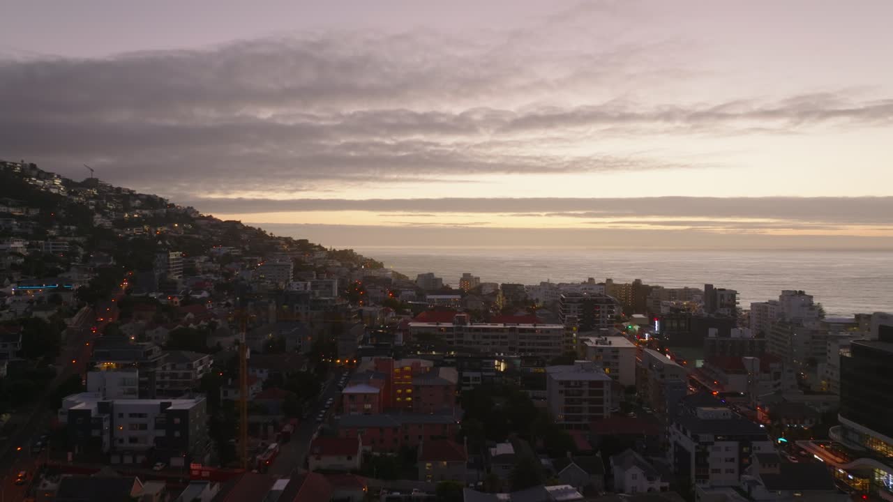 Vuelo hacia adelante sobre un barrio urbano costero al anochecer. Vista aérea contra un cielo de atardecer colorido con nubes. Ciudad del Cabo, Sudáfrica.