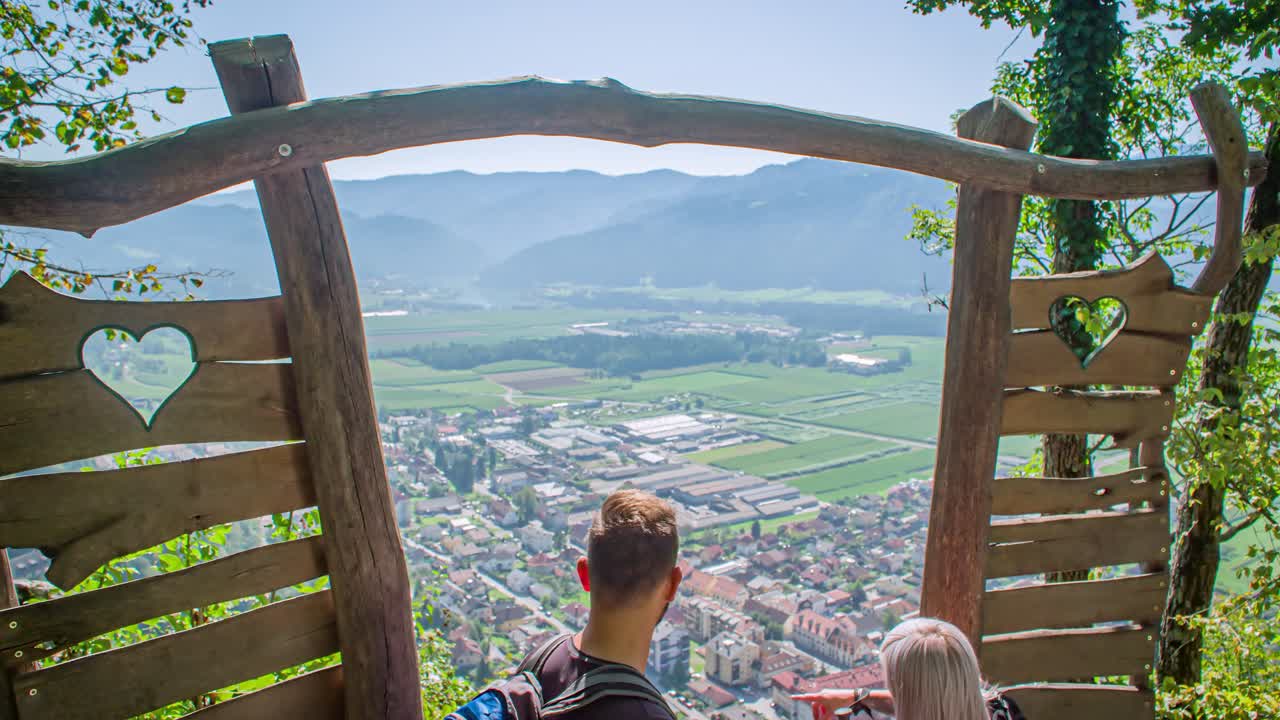 Panning shot up down. Couple looking out over a village and mountainous landscape. Green window, Slovenia