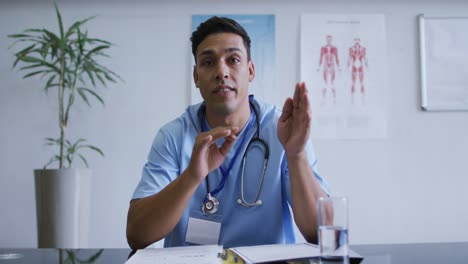 Mixed-race-male-doctor-at-desk-talking-and-gesturing-during-video-call-consultation