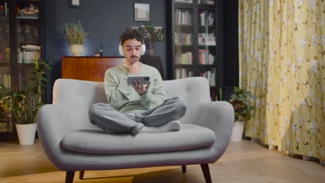 young man watching interesting movie on smartphone while sitting with crossed legs on couch at home