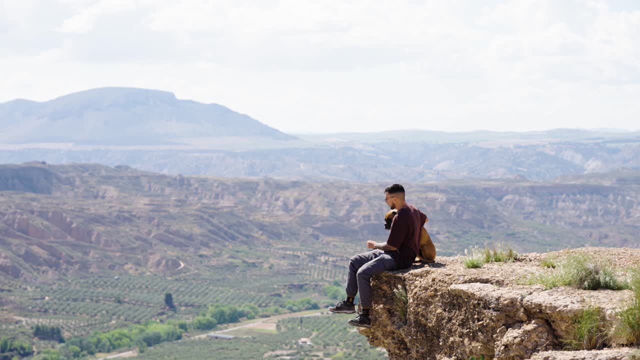 Man with Boxer dog sitting on high cliff overlooking valley landscape