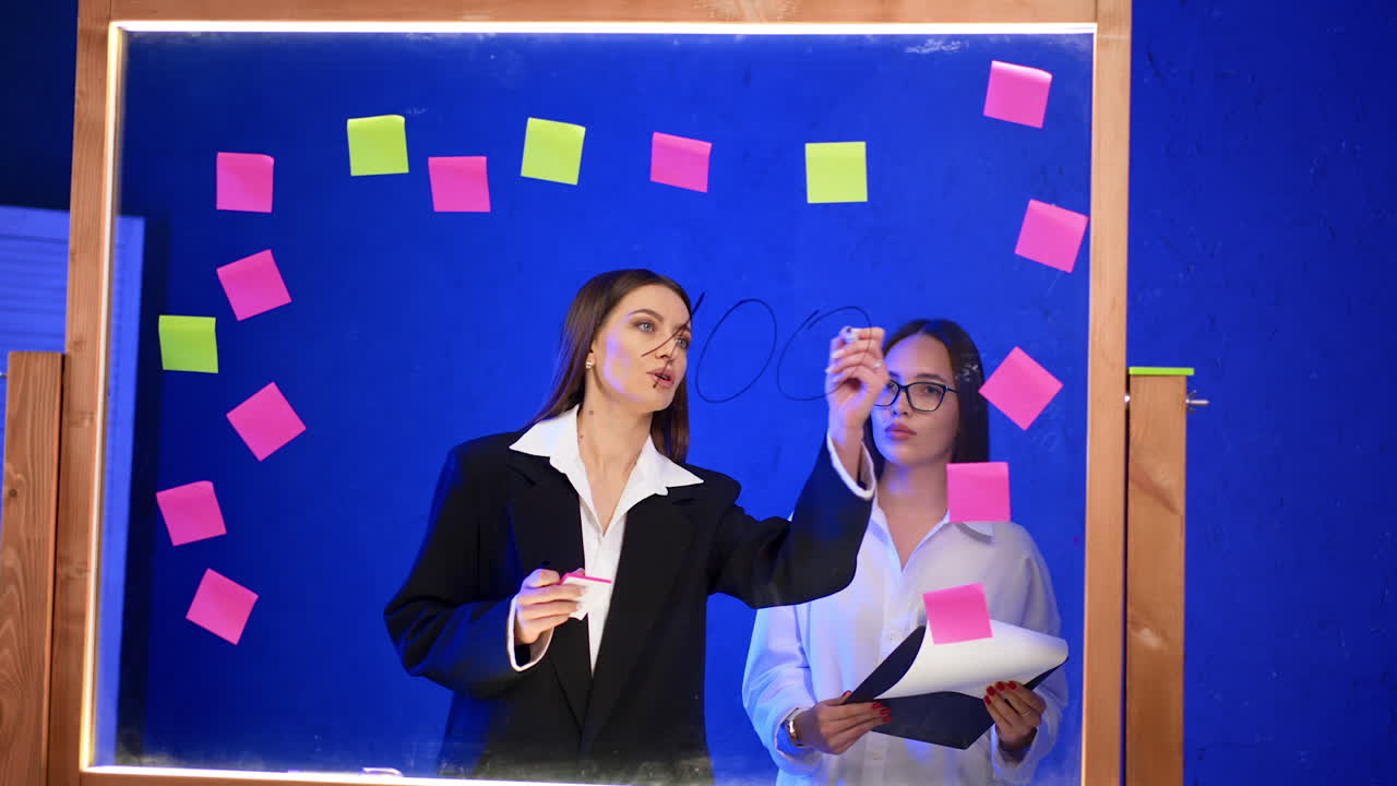 Ladies working in the modern office stand at the glass board. Woman in jacket writes percent numbers on the glass and woman in a shirt holds a clip board.