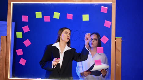 ladies working in the modern office stand at the glass board. woman in jacket writes percent numbers on the glass and woman in a shirt holds a clip board.