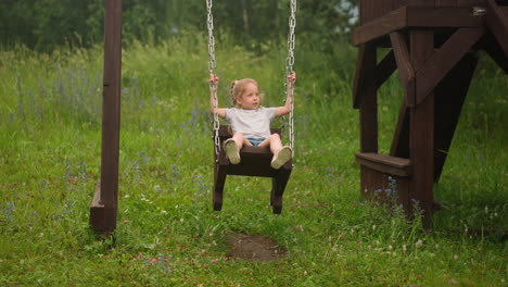 little girl plays swings on ground against high meadow grass