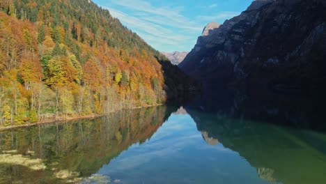 Vuelo-Aéreo-Sobre-Un-Tranquilo-Lago-Reflectante-Con-árboles-Forestales-Otoñales-En-La-Ladera-Del-Valle